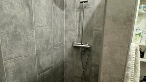 Une douche aux carreaux gris, un pommeau de douche à main et une serviette blanche posée sur un porte-serviettes.