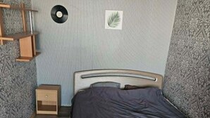 Une chambre avec un lit en bois, une table de chevet, un tourne-disque fixé au mur et une petite étagère.