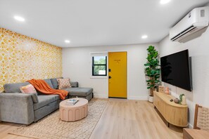Ein modernes Wohnzimmer mit einem grauen Sofa, einem runden Couchtisch, einer gelben Tür, einer großen Topfpflanze und einem an der Wand befestigten Fernseher.