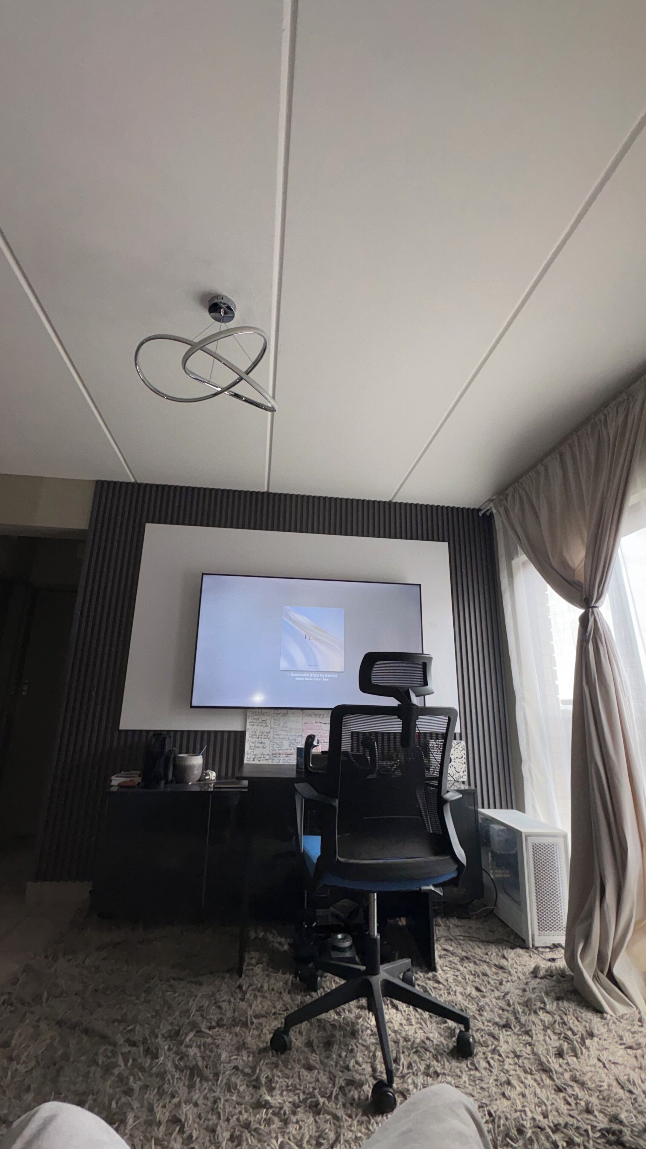 A hotel room with a large flat-screen TV mounted on the wall, a black office chair, a desk with a computer, and a radiator.