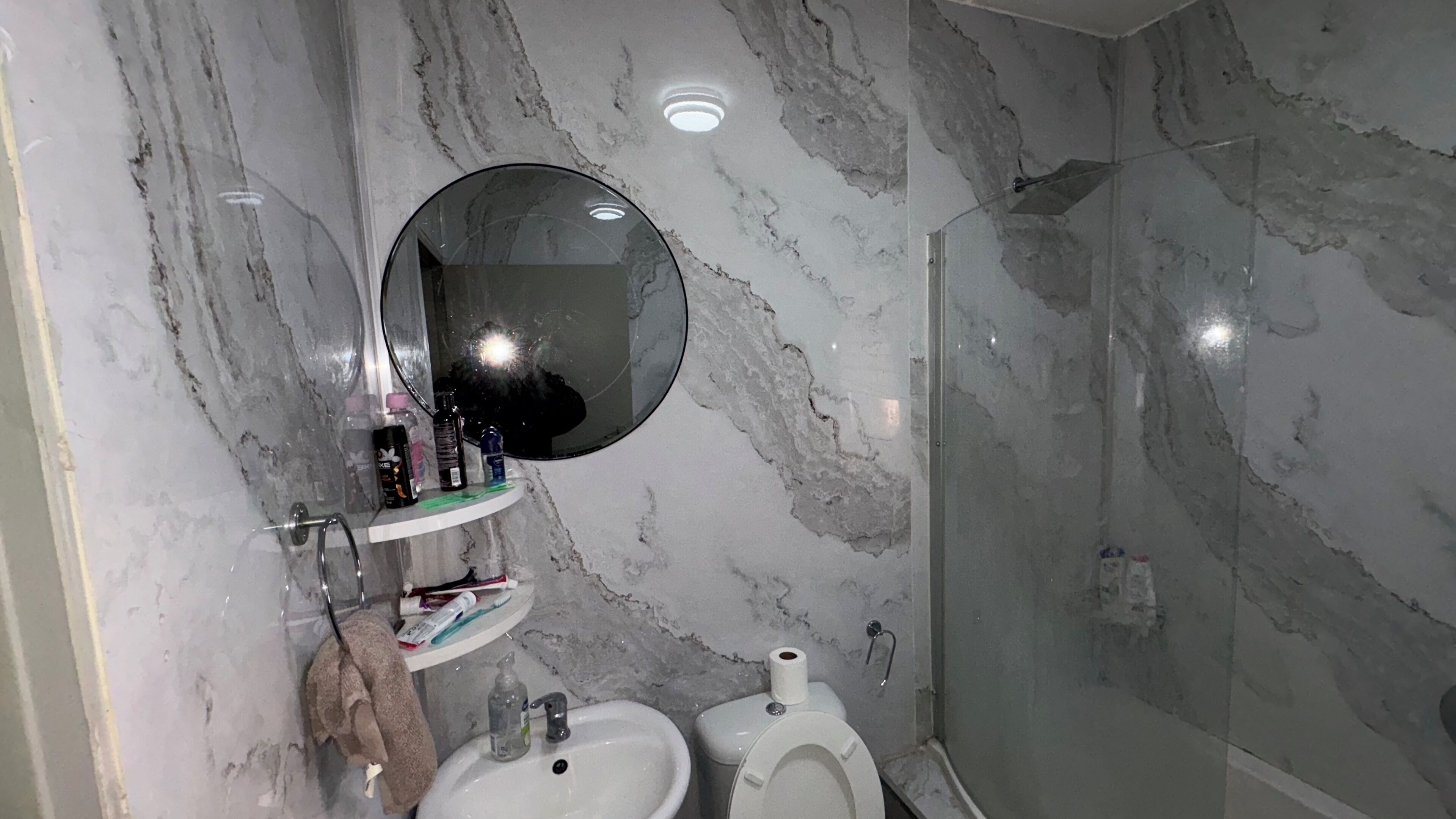 Un baño con pared de mármol, un espejo redondo, un lavabo, un inodoro y una cabina de ducha.
