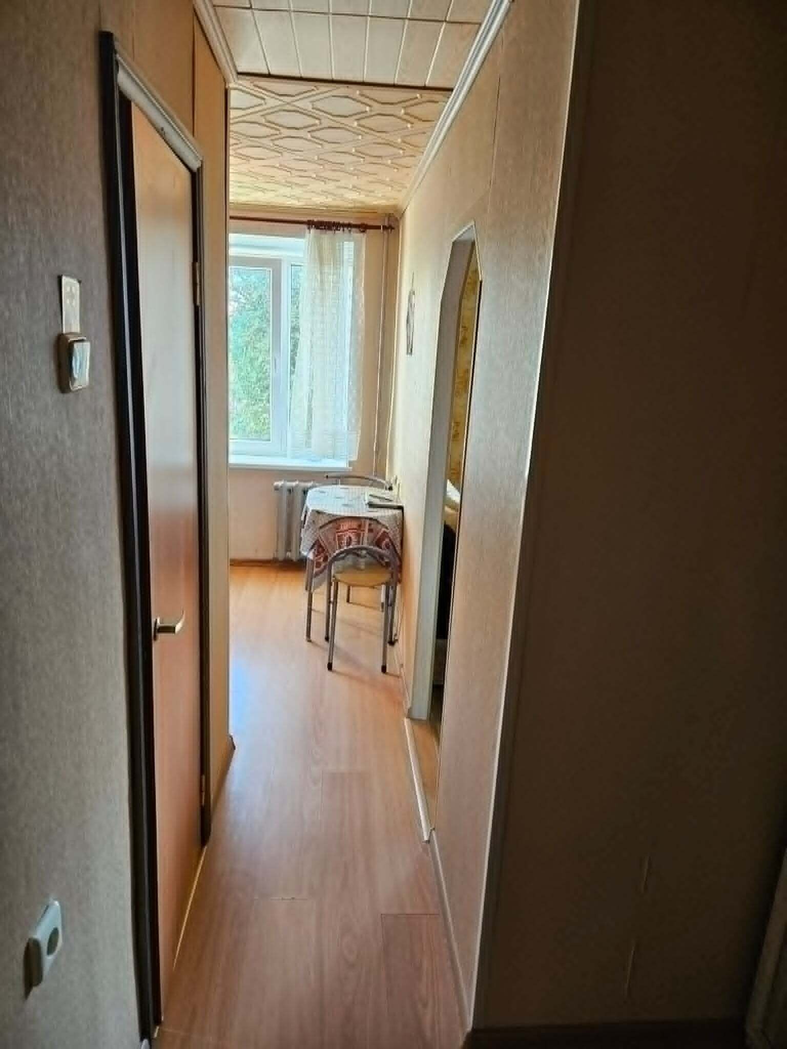 En smal hall med trägolv, ett element, ett bord med en stol och ett fönster med gardiner.