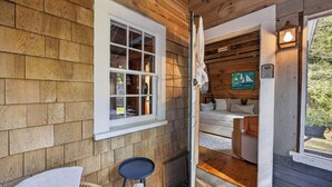 Interior de una cabaña de madera con una ventana, una puerta que da a un dormitorio, una mesita y una lámpara.