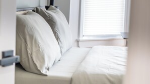 Een slaapkamer met een bed, kussens, een nachtkastje en een raam met jaloezieën.