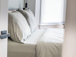 A bedroom with a bed, pillows, a bedside table, and a window with blinds. - Beautiful 2BR Home, Near NYC, PATH & Lincoln Park (Jersey City)