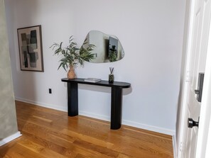 A hallway with a wooden floor, a black console table with a vase and a book, a mirror, and a framed abstract artwork on the wall. - Beautiful 2BR Home, Near NYC, PATH & Lincoln Park (Jersey City)