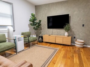 A living room with a sofa, a TV, a plant, a radiator, and a wooden cabinet. - Beautiful 2BR Home, Near NYC, PATH & Lincoln Park (Jersey City)