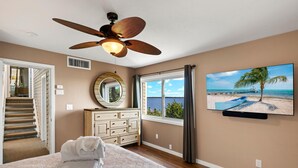 A bedroom with a bed, a ceiling fan, a dresser, a mirror, a window with a view of the ocean, and a television on the wall. - Widgeon Point (Fort Myers Beach)