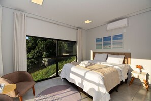 A modern bedroom with a large window, a bed with white linens, a bedside table with a vase of flowers, and a chair with a book. - Casa com Piscina Vista Mar e Banheira (Palhoça)