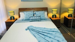 A neatly made bed with a blue blanket, two matching pillows, and two bedside lamps. A beach scene is visible on the wall above the bed. - Busselton Stylish 5 Bed, 3 Bathroom Home (Busselton)