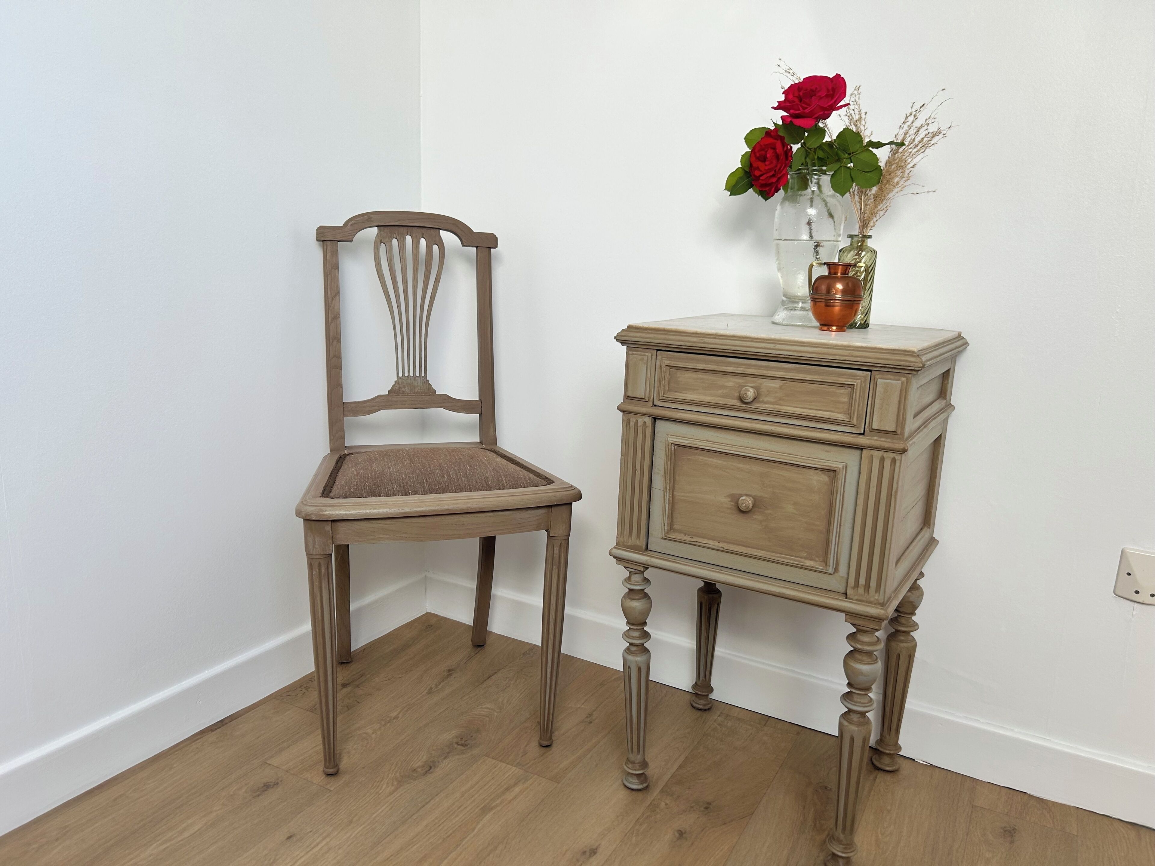 A wooden chair with a cushioned seat and backrest, a small wooden side table with drawers, and a vase with red flowers on the table.