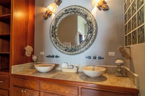 A bathroom with a wooden vanity, two large bowls, a decorative mirror, wall sconces, and a towel rack. - Experience Luxury by the Sea – Condo 16 at Oceans 23, Belize (San Pedro)