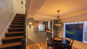 Una escalera conduce a un comedor con una mesa y sillas, una cocina al fondo y un amplio ventanal con vistas.