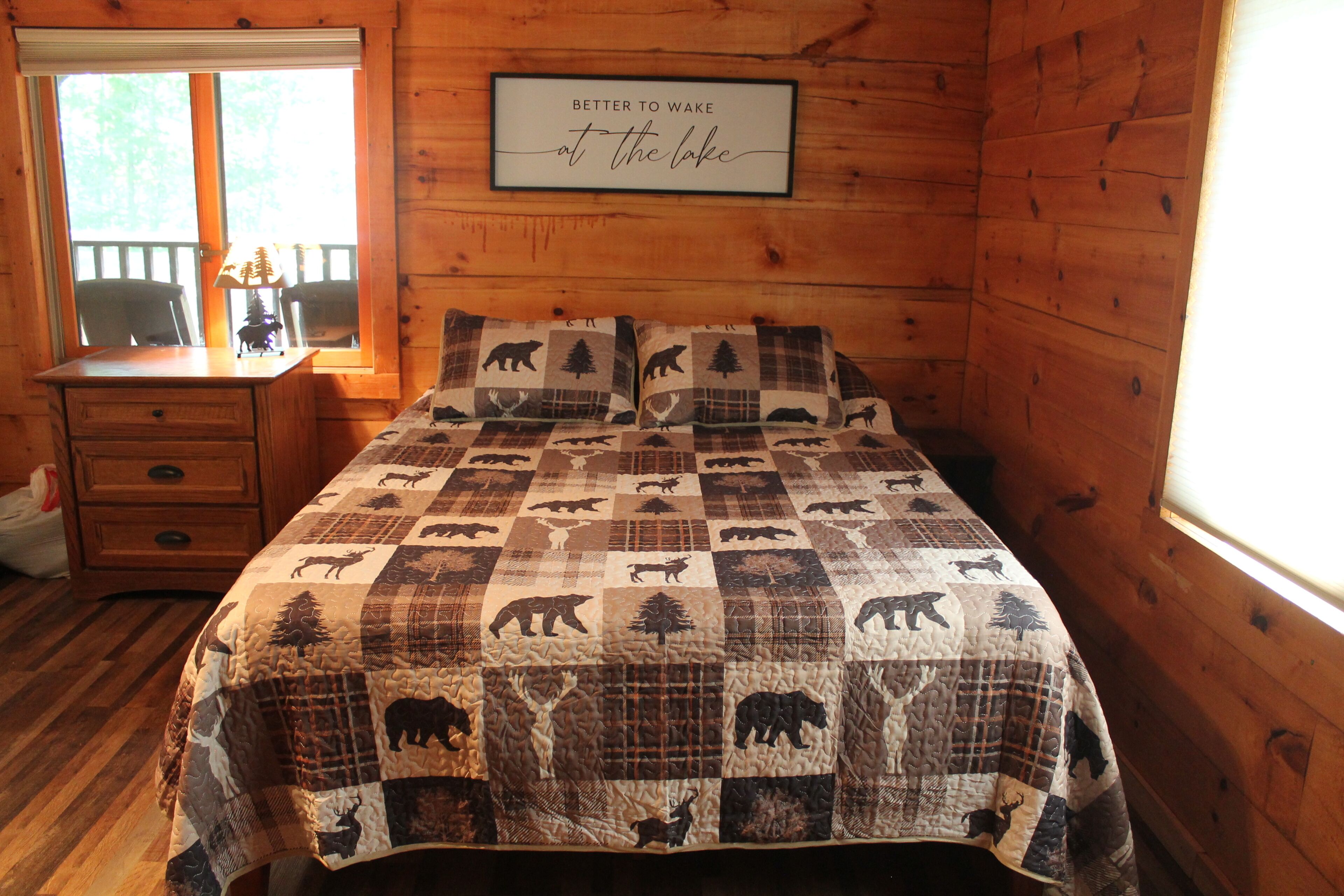 Une chambre dans un chalet en bois, avec un couvre-lit matelassé orné de motifs animaliers, une commode en bois, une fenêtre avec des stores et une citation encadrée accrochée au mur.