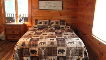Une chambre dans un chalet en bois, avec un couvre-lit matelassé orné de motifs animaliers, une commode en bois, une fenêtre avec des stores et une citation encadrée accrochée au mur.