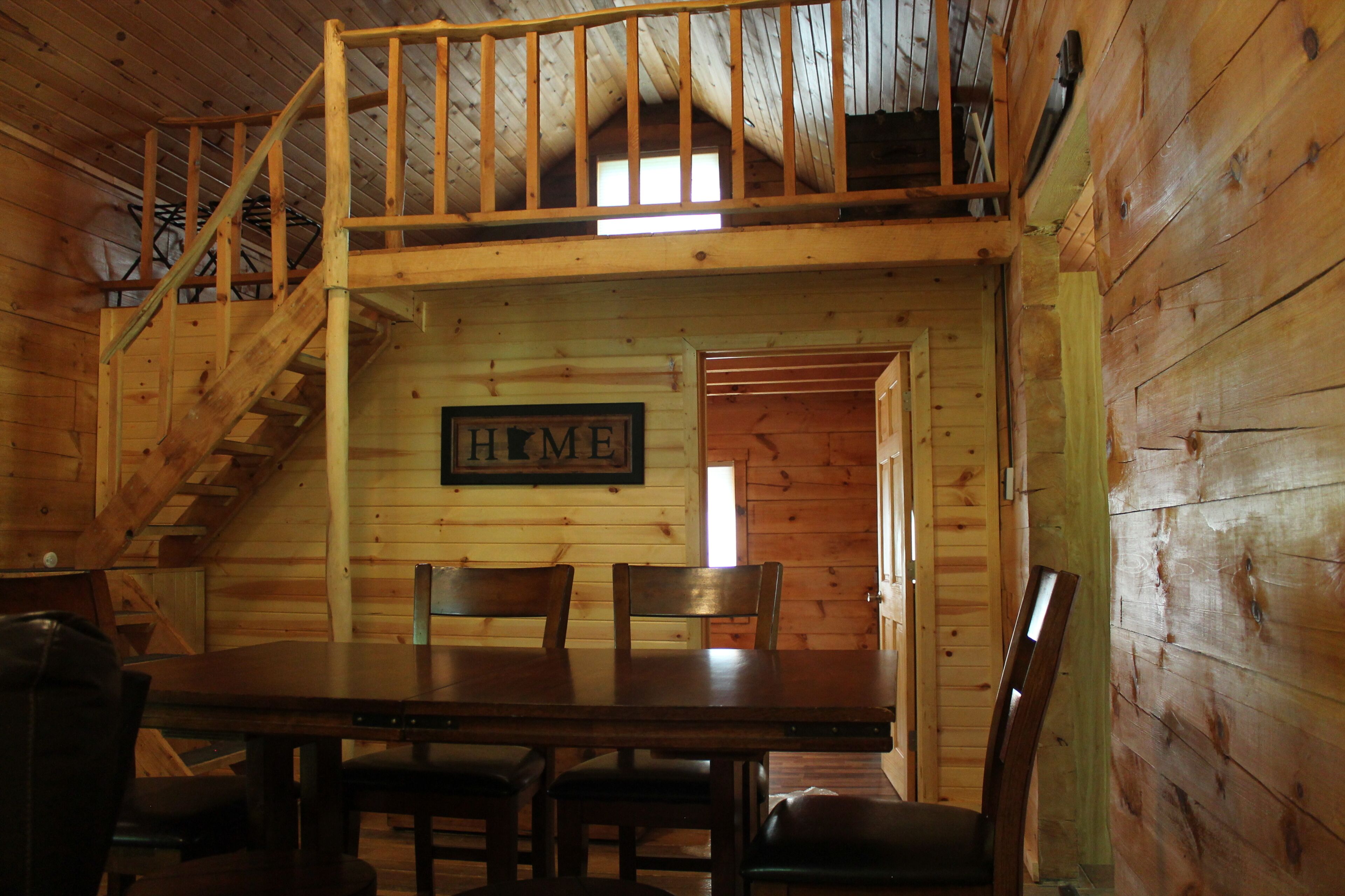 L’intérieur d’une cabane en bois, avec une table à manger, des chaises, un escalier et un panneau indiquant « MAISON » sur le mur.