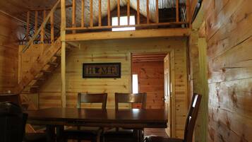 L’intérieur d’une cabane en bois, avec une table à manger, des chaises, un escalier et un panneau indiquant « MAISON » sur le mur.