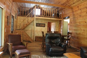 A wooden cabin interior with a loft bedroom, a dining area, and a living room furnished with leather chairs and a sofa.