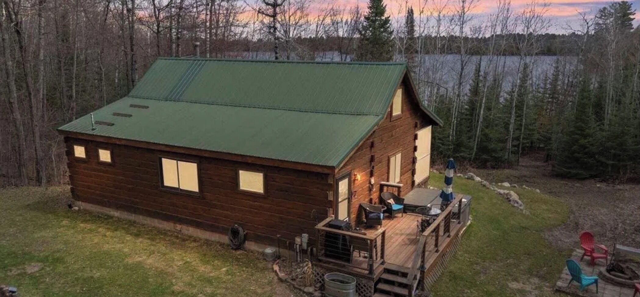 Une cabane en bois avec un toit vert, une terrasse dotée de meubles d’extérieur et une vue sur une zone boisée.