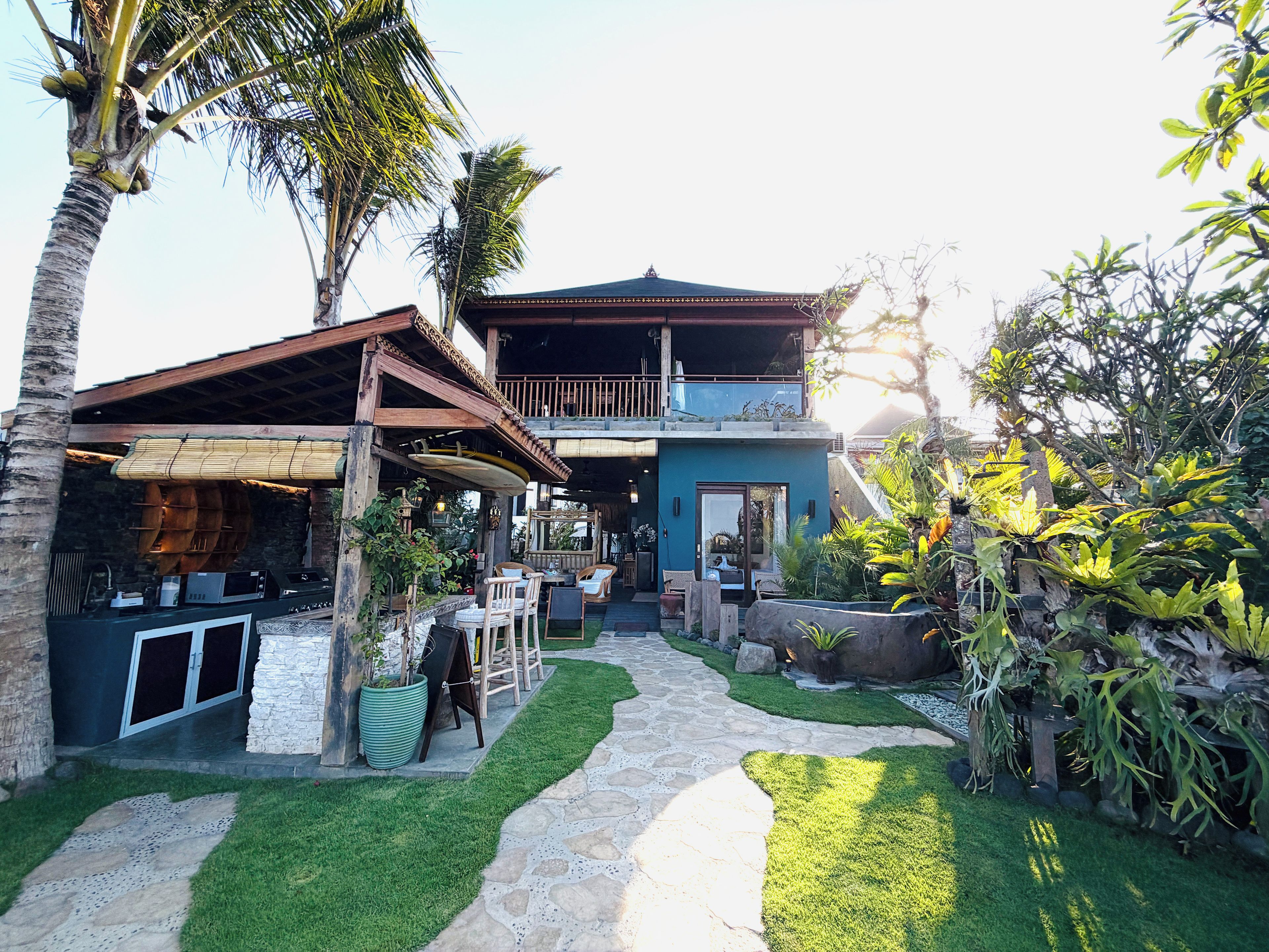 A two-story building with a balcony, surrounded by a well-maintained garden with palm trees and various plants.