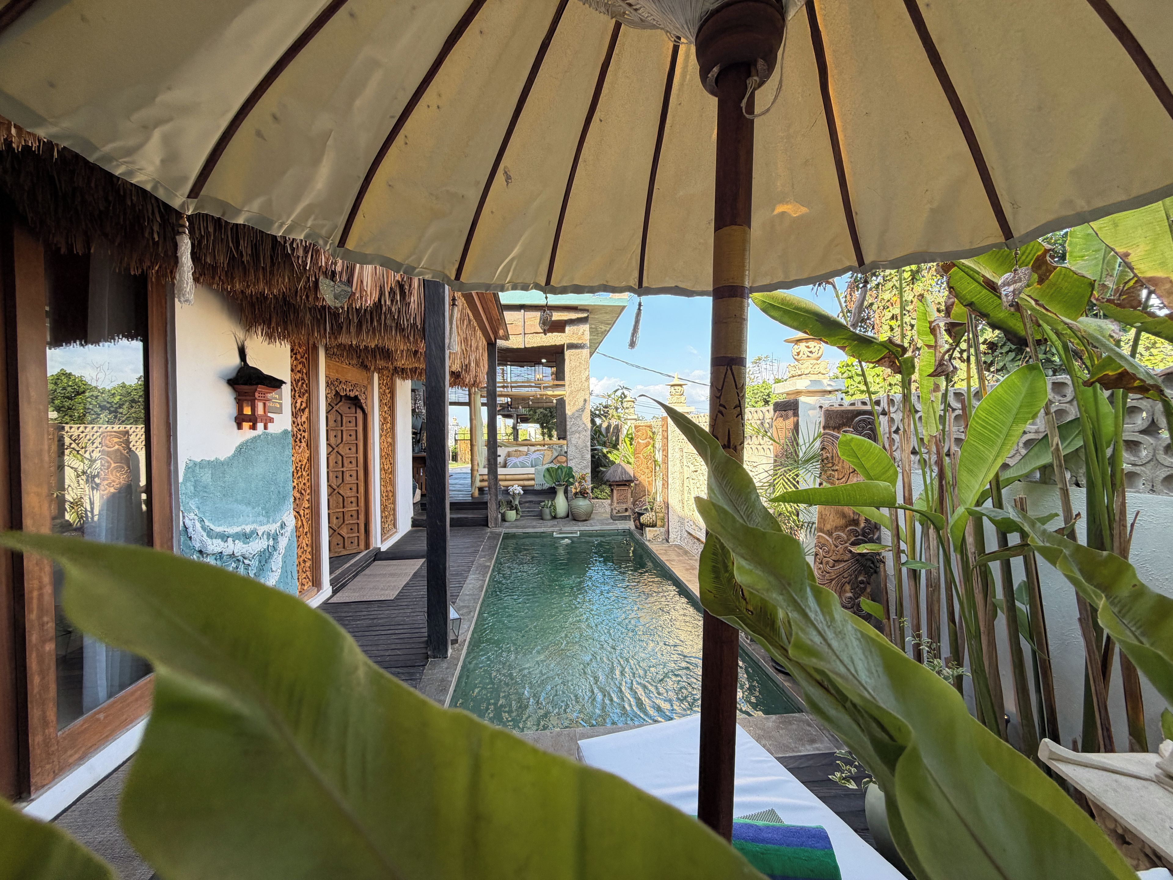 Un área de piscina con una estructura de techo de paja, una terraza de madera y plantas tropicales.