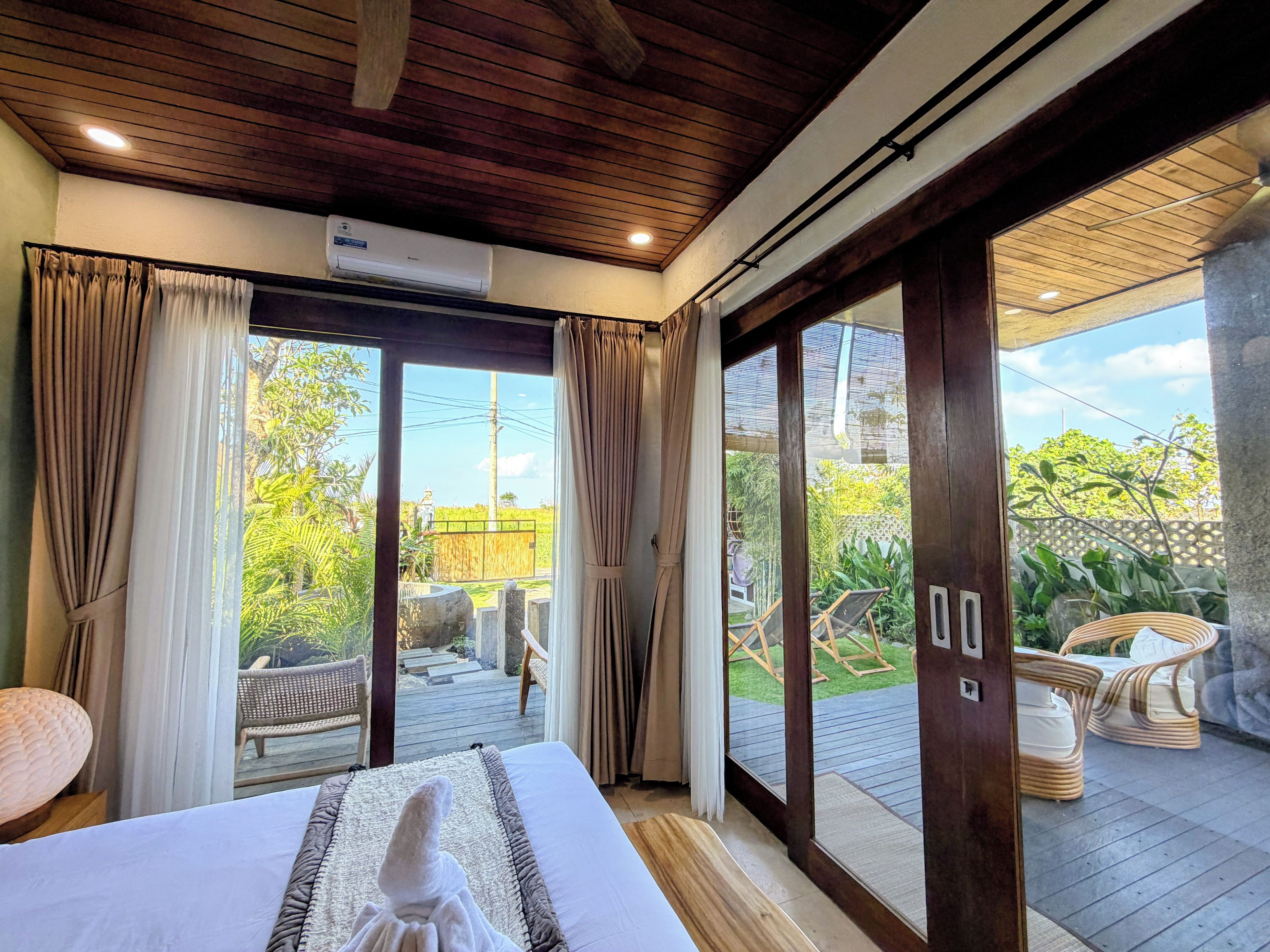 Une chambre d’hôtel avec un lit, des rideaux, un appareil de climatisation et une vue sur la terrasse extérieure, qui est aménagée avec du mobilier.