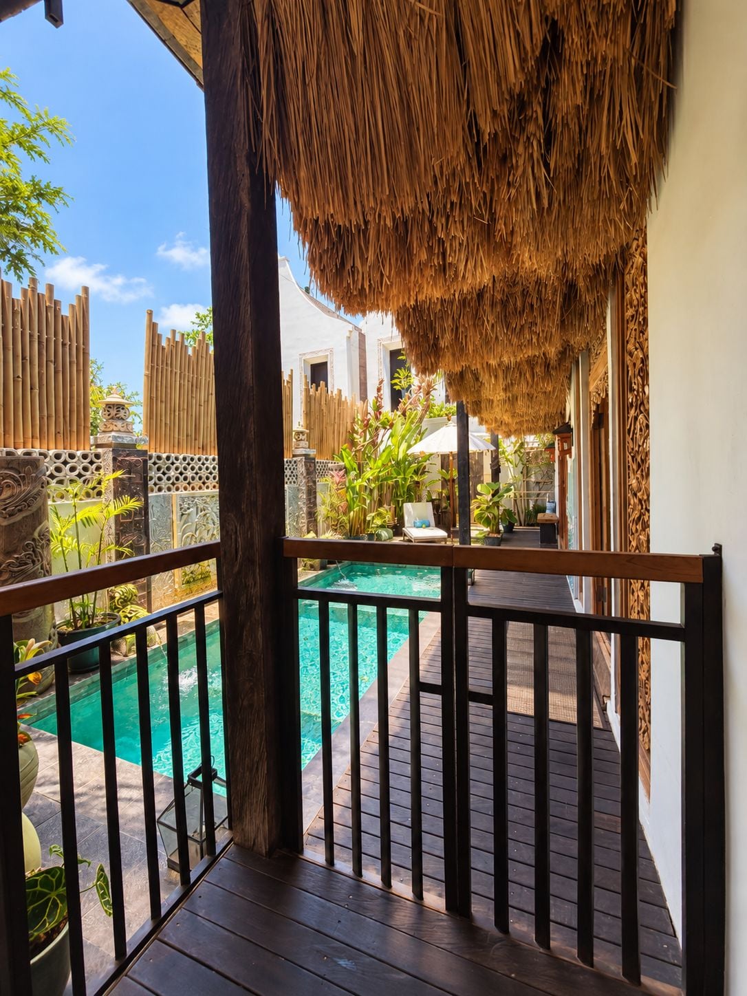 A wooden deck with a thatched roof, overlooking a swimming pool and a garden area with plants and a white building in the background.