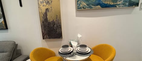 A dining area with two yellow chairs, a small round table, and a painting on the wall.