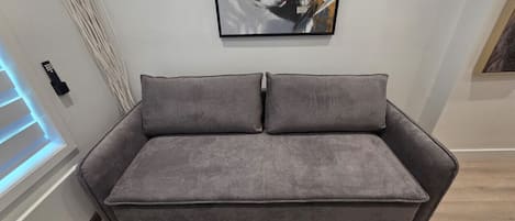A grey sofa with two cushions in a room with a modern design, a painting on the wall, and a window with blinds.
