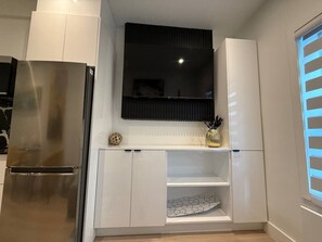 A modern kitchen with white cabinetry, a stainless steel refrigerator, a black TV mounted above a white shelf, and a window with blinds. - Downtown like no other! (Montréal)