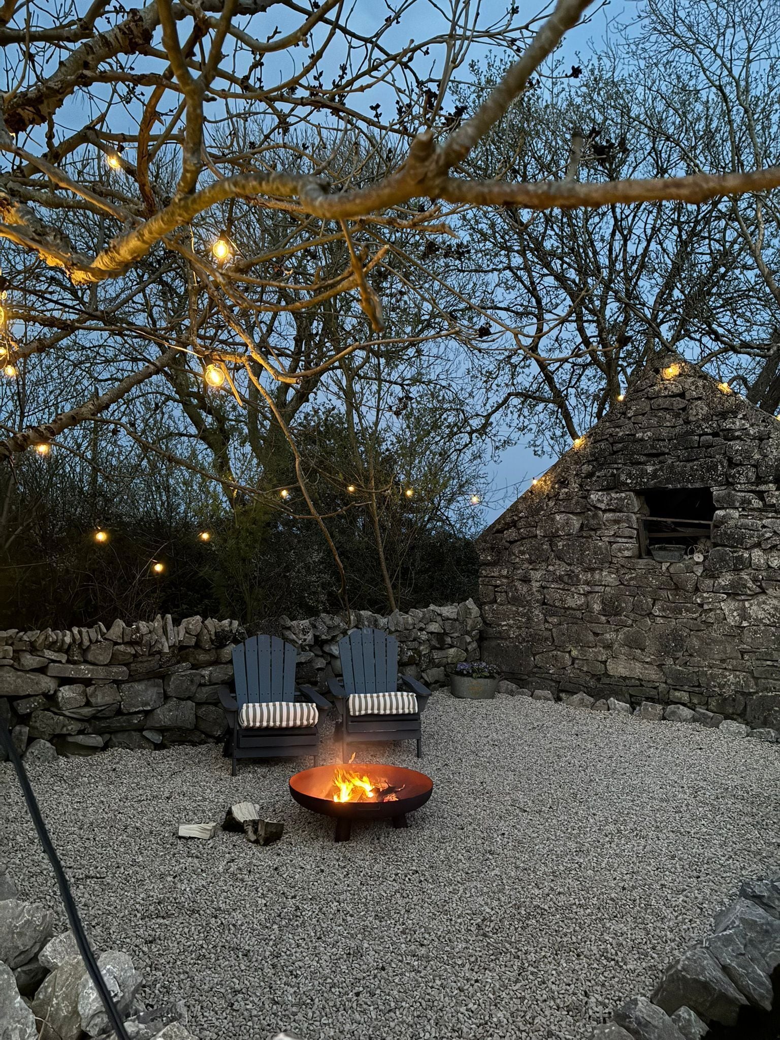 Kivimuuri, kaksi Adirondack-tyylistä puutarhatuolia, nuotiopaikka ja valonauhat koristavat puutarhaa.