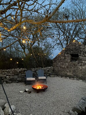 A stone wall, two Adirondack chairs, a fire pit, and string lights in a garden setting.