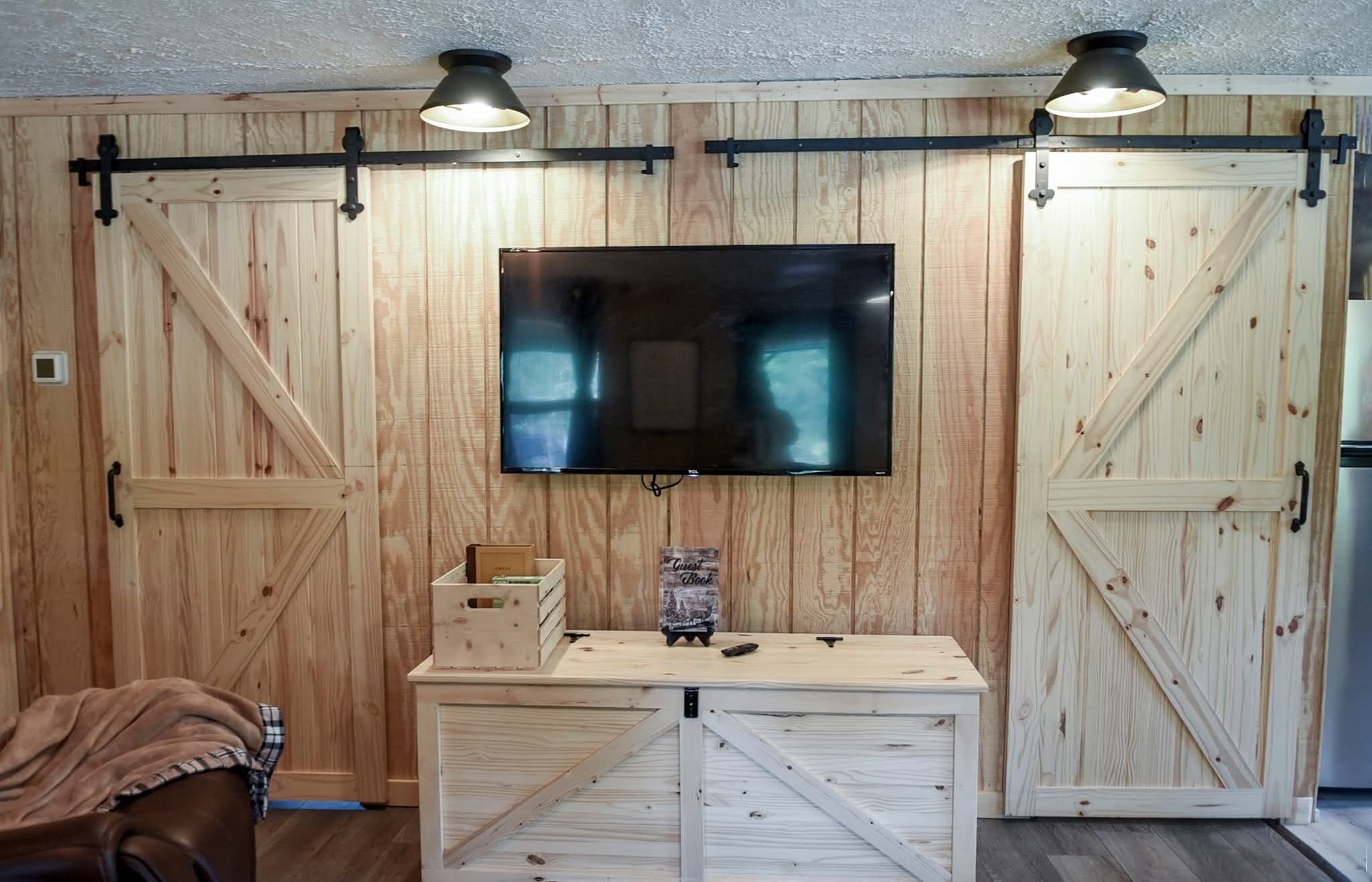 A room with wooden walls, a mounted flat-screen TV, a wooden table with a box and a remote, and a couch with a blanket.