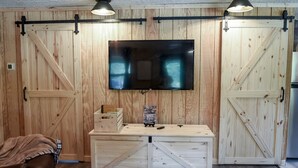 Una habitación con paredes de madera, un televisor de pantalla plana montado, una mesa de madera con una caja y un control remoto, y un sofá con una manta.