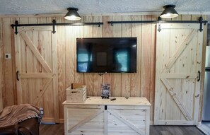 A room with wooden walls, a mounted flat-screen TV, a wooden table with a box and a remote, and a couch with a blanket. - LakeEffectLodging-Cozy Cabin-ORV Trail Access (Luther)