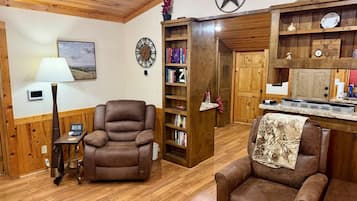 A room with a wooden ceiling, a fireplace, a bookshelf, a clock, and a dining area.