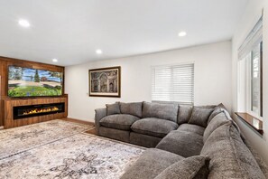 Ein Wohnzimmer mit Flachbildfernseher, Kamin, grauem Sofa, gemustertem Teppich und einem gerahmten Bild an der Wand.