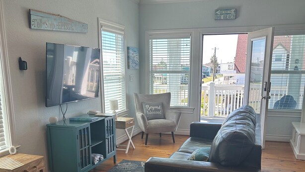 Ein Wohnzimmer mit einem schwarzen Ledersofa, einem türkisfarbenen Beistelltisch, einem Flachbildfernseher und Blick auf den Strand durch große Fenster.