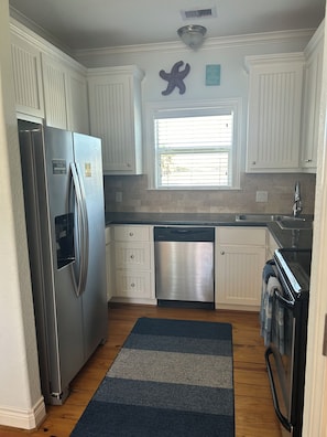 A kitchen with white cabinets, a stainless steel refrigerator, a dishwasher, a window with blinds, and a starfish decoration. - Cozy 2 BR/1.5BA near West End Marina in Sea Isle!  https://seaisleperch.com/ (Galveston)