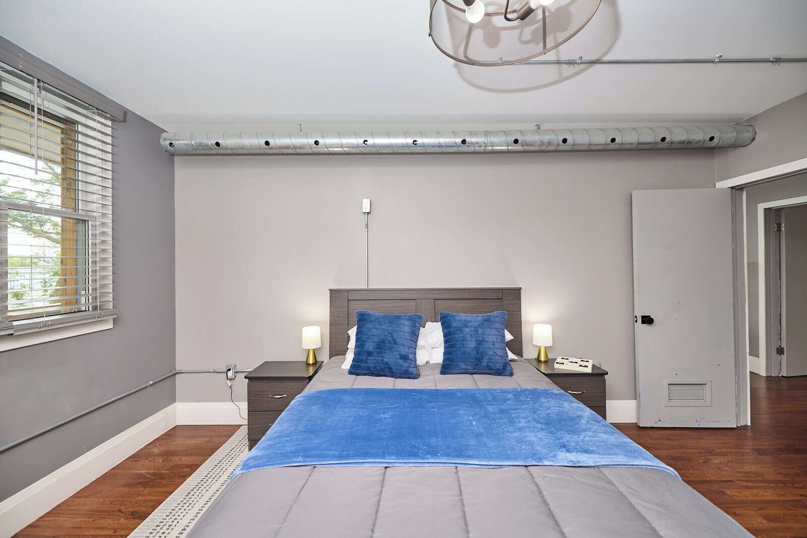 A bedroom with a bed, two bedside tables, a nightstand, a window with blinds, a door, and a ceiling light fixture.