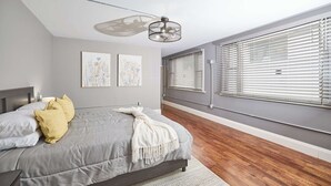 A bedroom with a bed, a ceiling fan, artwork on the wall, and large windows with blinds.
