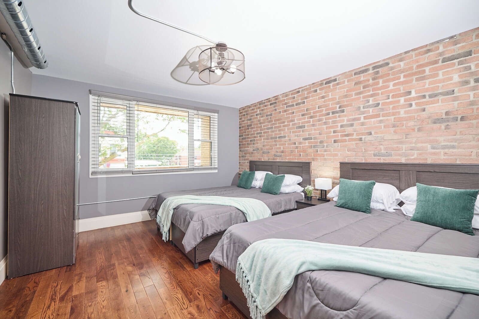 Une chambre avec deux lits, un mur en briques, une fenêtre avec des stores et un ventilateur de plafond.