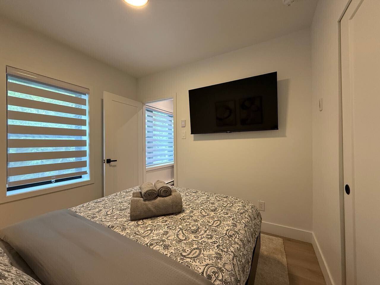 A bedroom with a bed, two windows with blinds, a flat-screen TV mounted on the wall, and a door.