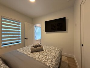 Une chambre à coucher avec un lit, deux fenêtres équipées de stores, un téléviseur à écran plat fixé au mur et une porte.