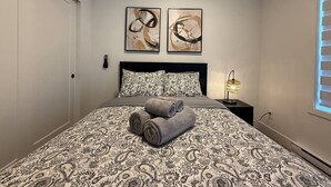 A bedroom with a patterned bedspread, two abstract paintings above the bed, a bedside lamp, and a window with blinds.