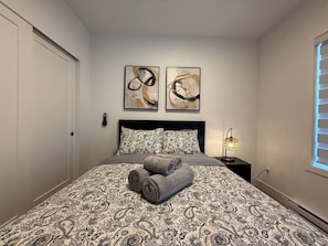 Un dormitorio con una colcha estampada, dos cuadros abstractos sobre la cama, una lámpara de noche y una ventana con persianas.