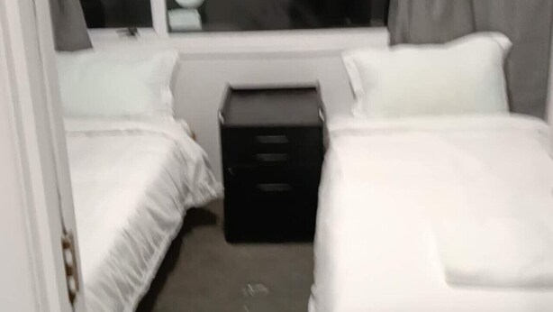 A hotel room with two single beds, a small black nightstand, a window with curtains, and a view of the outside. - 2 Bedroom Garden Chalet (Rotorua)
