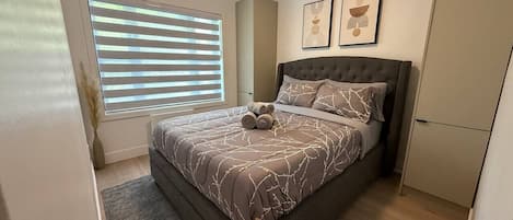 A bedroom with a bed, a window with blinds, a bedside table, and a wall-mounted artwork.