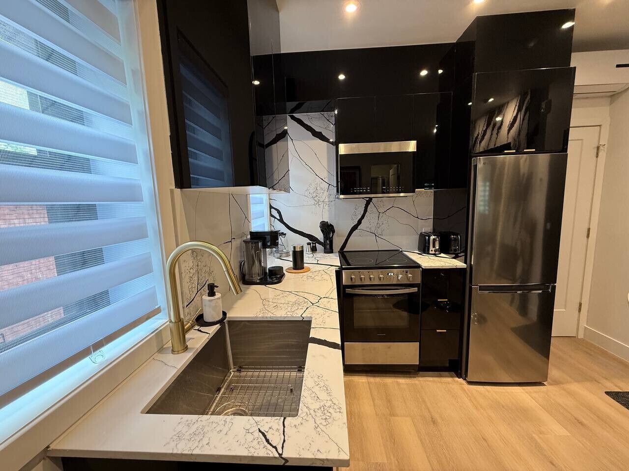 A modern kitchen with a built-in oven, a stainless steel refrigerator, and a window with blinds.