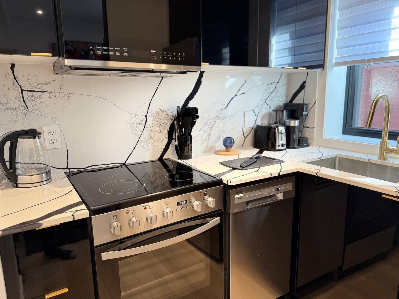 A modern kitchen with a built-in stove, oven, and dishwasher, a marble backsplash, and a window with blinds.